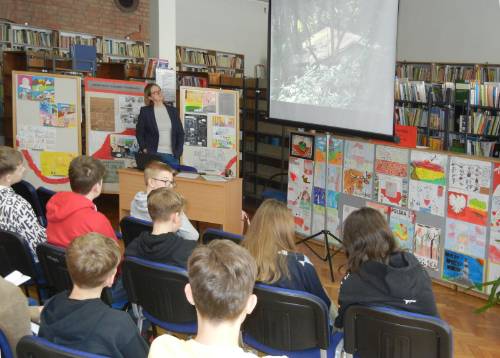 Widok od tyłu na młodzież uczestniczącą w zajęciach w bibliotece, w tle widać prowadzącą dr Barbarę Męczykowską oraz ekran z prezentacją i rysunki ozdabiające ściankę.